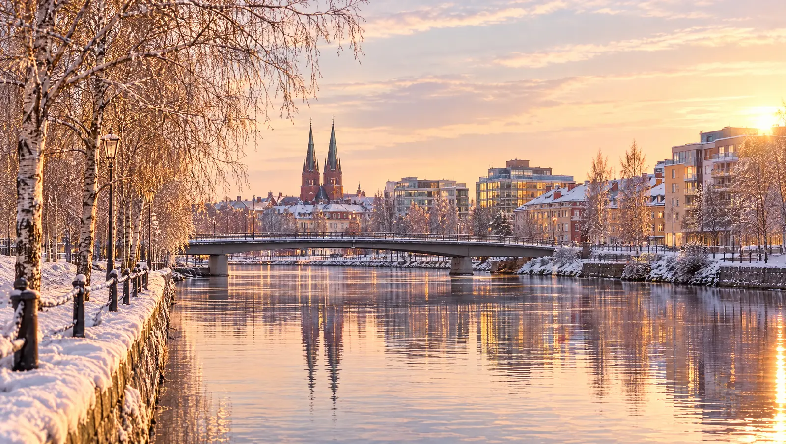 Stadssiluett vid en bred älv en kall morgon, med den första snön på kajen och gula löv kvar i träden